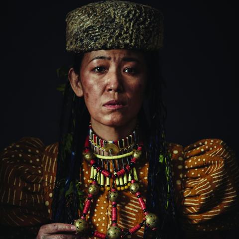 A women in a thick beaded necklace and a flat hat stares solemnly