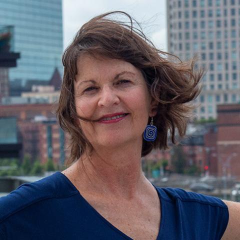 Cheri is a white woman with jaw-length brown hair. She wears a navy v-neck and poses in front of city buildings.