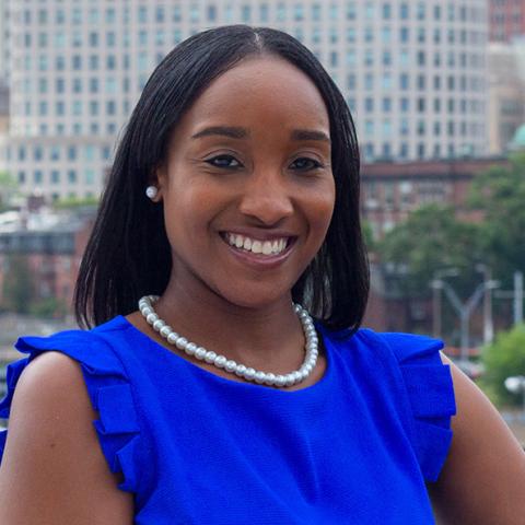 Indira is a Black woman in a bright blue blouse. She poses in front of city buildings.