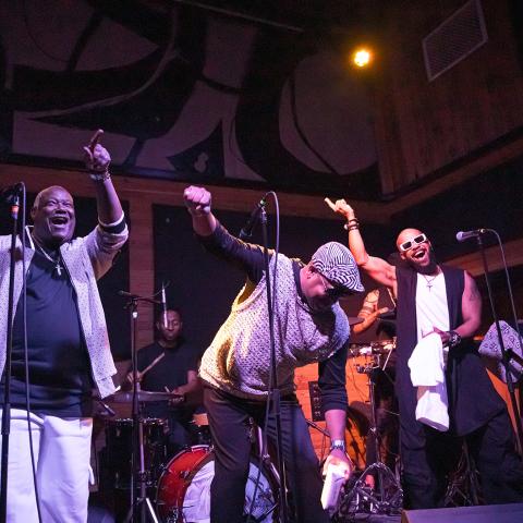 Three Black men dance and point. One bends over. A band is behind them. 