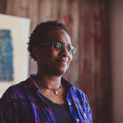 Chelvanaya is a Black person. They wear a flannel shirt with a deep v t-shirt underneath. They have thin-framed glasses. A work of art hangs on the wall behind them.