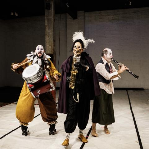 On a stage, three men in clown gear that is in sad hues and one of them has a skull mask on..