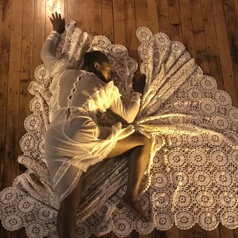 On a wood floor at sunset, a Black woman in a white lace dress lays on a white lace blanket.