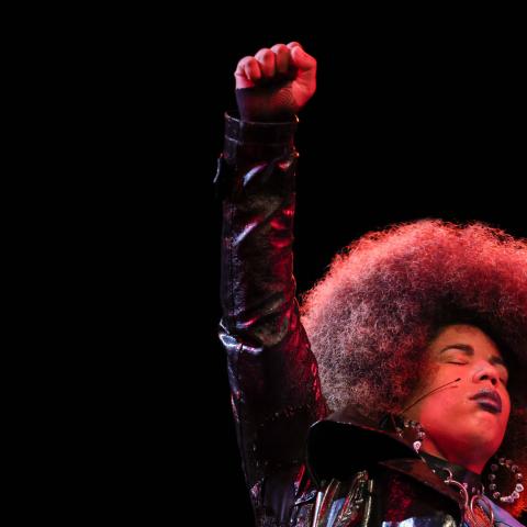 A Black girl, with an Afro, holds up her fist in a spotlight.