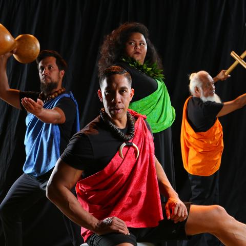 In front of a black curtain, four folks in different colored satin sashes pose. Two pose with their drum or drumsticks.