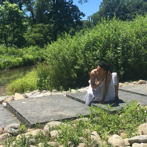 Along a stone path, a woman of color, dressed in white, crouches on one knee.