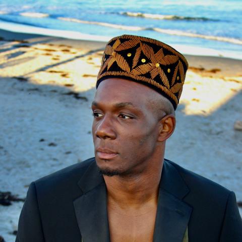 Harold is a Black man in a kufi and a black blazer. They are on the beach with the water hitting the coast behind them.