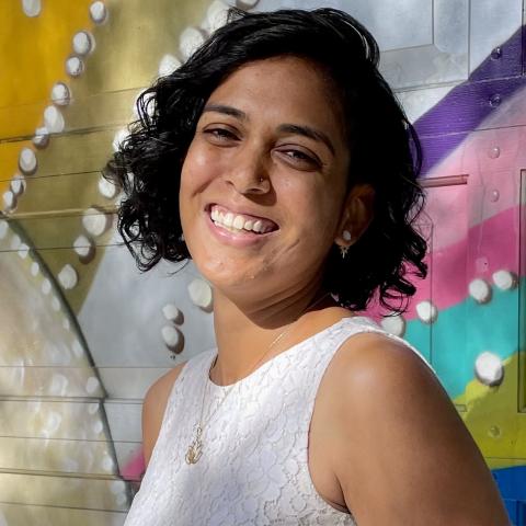 Heena is a brown woman with jaw-length wavy black hair. She wears a whtie tank top in front of a colorful mural.