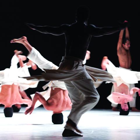 male dancers wearing white pants; several appear to be twirling on their heads while others are standing up with arms stretched.