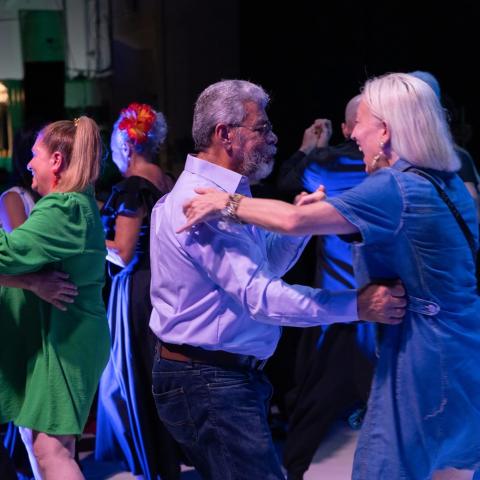 In the foreground, an elderly heterosexual couple dances. Behind them, younger couples of various combinations dance.