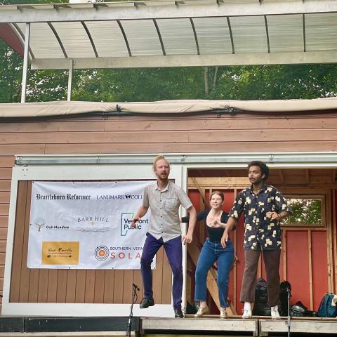 Three performers on a small outdoor stage with a wooden facade and sponsor banner.