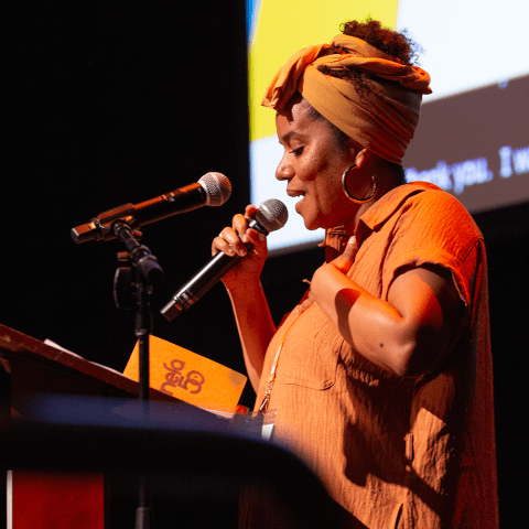 A brown woman speaks into a microphone. She wears an orange blouse and headwrap. She holds her chest.