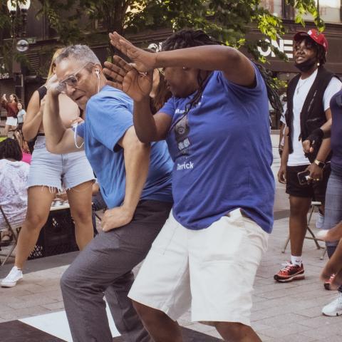 Outside, at a downtown shopping center, two folks lean into each other while dancing. Other folks of different backgrounds and identities watch and dance too.