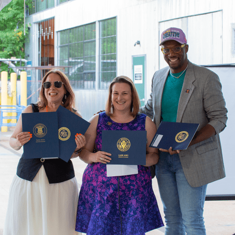 A white woman, in sunglasses, poses with another white woman, in a purple and blue dress, and a Black man in a grey blazer and light purple hat.