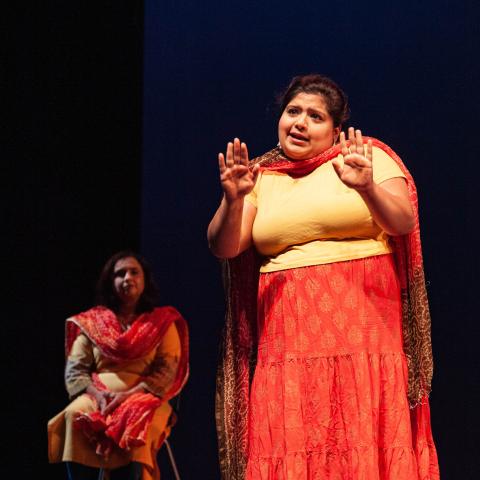 Two brown women are on stage. They wear red and yellow outfits, including wraps that lay by their necks and across their bodies. The woman in the foreground holds her hands up.