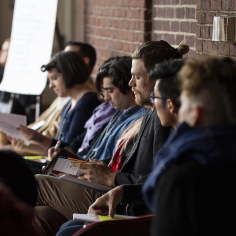 Indoors, a row of people sit and look at booklets they're holding.