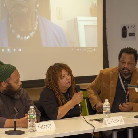 Three people sit behind a table with microphones on it. In front of them there are handwritten name tags, "Kenny," "L'Merchie," and "Steve."