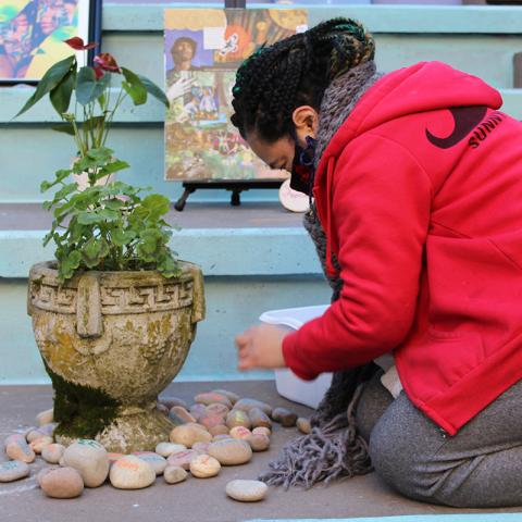 A woman, in red sweatshirts,  places a piece of paper next to a plant, in a stone vase.
