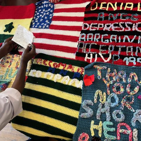 Black hands hold a piece of paper of a knitted quilt with blocks that feature the American flag, "Denial, Anger, Depression, Bargaining, Acceptance, HIV 1999," "Thank you North Shore Arts," and a yellow and black square.