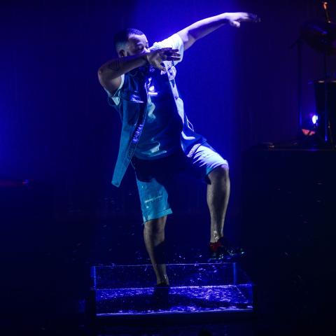 On a stage, a Black man lifts his leg and raises both arms to the right and across his face.