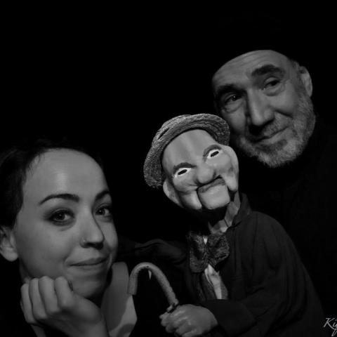 In black and white, a young white woman and an older white man, in a black beanie that fades into the background, pose with a wooden puppet.