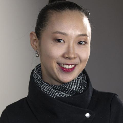 A woman wearing a black funnel-collard jacket and silver drop earrings. Her hair is pulled back into a bun.