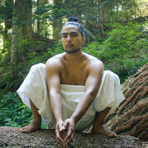 In the woods, a shirtless man leans over a fallen tree, posing for the camera.