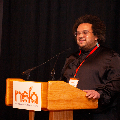 A person stands behind a podium. They wear a satin shirt and a nametag with an orange lanyard.