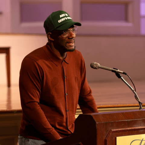 A Black man, in a trucker hat and burnt orange blouse, stands behind a podium.