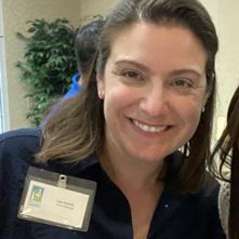 Lee has shoulder length brown hair. She smiles and wears her nametag. She's with three other women but the image is sometimes cropped to just include her face.