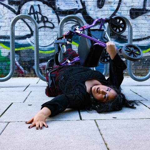 a photo of Vanessa, who is a Chicana woman with caramel skin is lying down on the concrete floor with her arm reaching out as she is looking back towards the camera. Her walker Pluto is balancing on top of her. She is wearing a gothic long sleeve blouse with a flowered black and red skirt. In the background is a looping metal bicycle stand with a graffiti filled wall.]