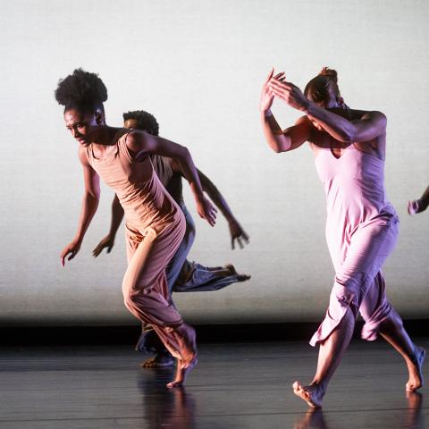 Four Black dancers, on a stage and in front of a white backdrop, lean forward with their left rear foot raised and their arms up or at their sides.