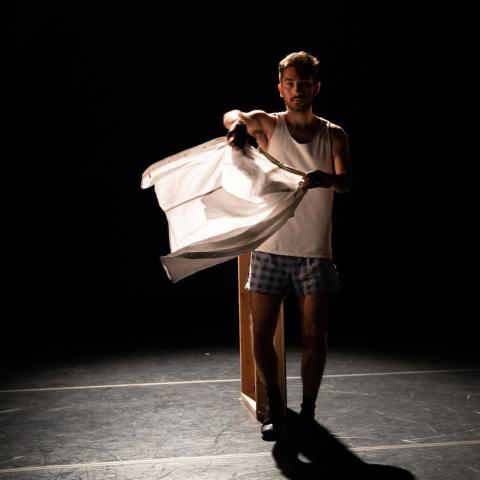 A man stands in a darkened black box theater in an undershirt and boxers waving a white tee shirt like a flag