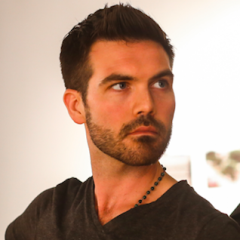 John looks to the side while he wears a beaded necklace and a v neck t-shirt.