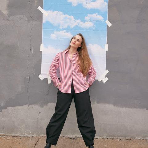 A white, blonde lady poses in front of a small backdrop of clouds that has been taped to the wall.