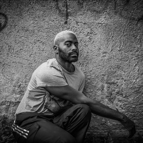 Black and white photo of Jerron as he crouches against a cracked brick wall, both arms laying to his side.