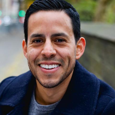 A head shot of a man smiling, wearing a navy overcoat and sitting outside.