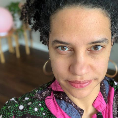 Jess Roseman makes eye contact in front of plants and a globe lamp, in a pink and multicolor patterned top. 