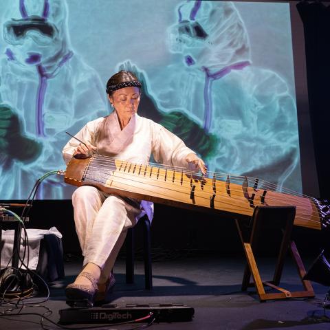 Jin Hi plays the electric komungo, wearing all white in front of a projection of folks in hazmat suits.