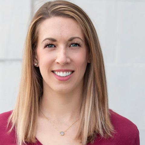 A white woman with medium length straight blonde hair wearing a v-neck red top and subtle jewelry.