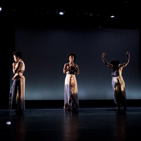 On an indoor stage, five Black, female dancers perform in a line. They wear monochrome gowns.