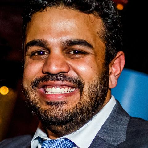 A brown man, in a suit and with curly dark hair, grins.