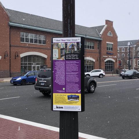 Image of a sign on a lamp post on a sidewalk with the town center in the background.