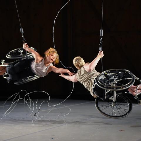 Alice, a dancer with light brown skin and short curly hair, and Laurel, a dancer with pale skin and close-cropped blonde hair, hang from the ceiling. They each have one hand on a cable as they reach for each other with the other. They are nearly horizontal and their legs and feet face out. Silver barbed wire stretches between and beneath them, against the black backdrop.