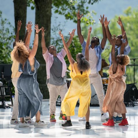 At an outdoor space, six Black dancers, in jumpsuits and khakis, hold their hands up to the sky.