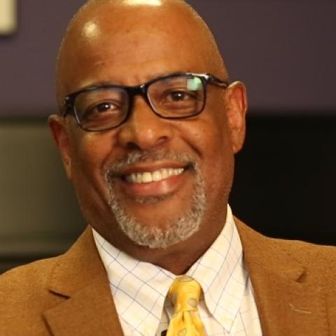 A man with glasses and a short beard wearing a yellow tie, white shirt, and caramel blazer, sitting in an office setting.