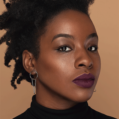 A Black woman with short hair wearing  a black top and bold silver earrings. She is in front of a neutral background.