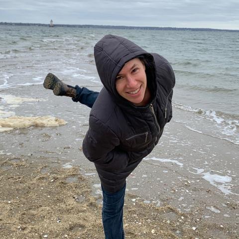 A white woman wearing a hoody standing n arabesque on a beach on an overcast day.