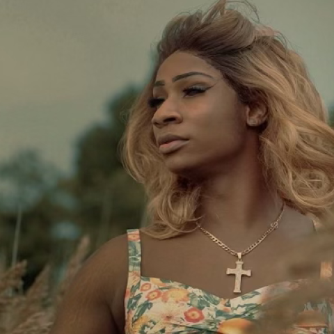 A Black woman standing in a field looking into the wind. She wears a floral printed top and a large gold cross around her neck.
