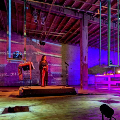 A woman, in a wrap dress, stands on a stage next to a music stand. The space has purple lights and projection of a similar fabric. It's empty otherwise.